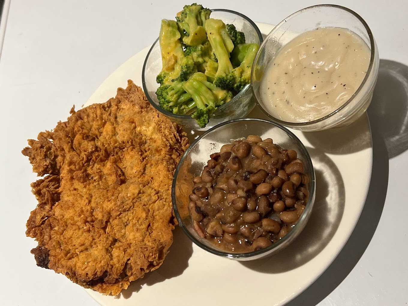 A white plate with fried chicken, and three glass bowls filled with beans, broccoli, and gravy.