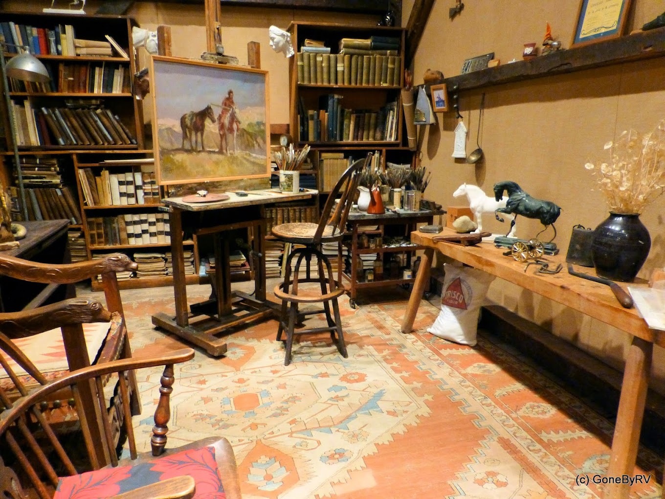 Room with bookshelves filled with books, painting stand with a chair, and sculptures.