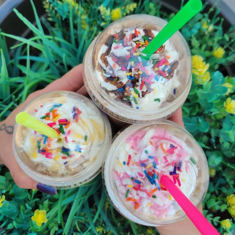 An overview of three drinks with whipped cream and colorful sprinkles topping the drinks, on a background of grass and flowers.