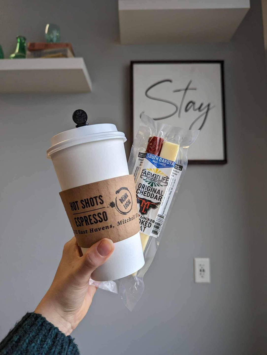 Someone holding a latte in a white to-go cup with a brown paper in the middle with a logo, a meat and cheese stick in a package is held in the same hand with the coffee.