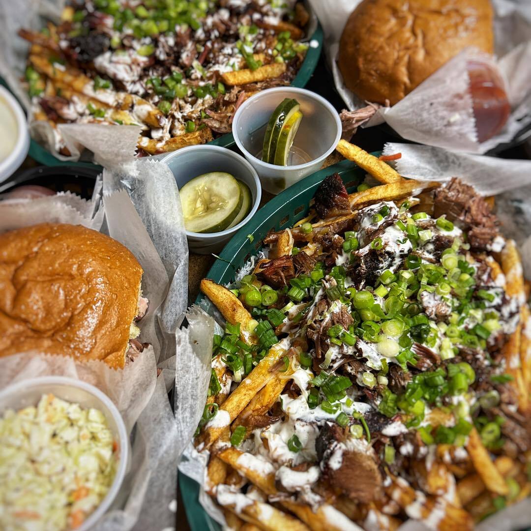 Table filled with different dishes. There are two plates of steak, fries, green onions, and sauce; there are two plates of burgers. There is a cup of coleslaw and two cups of cucumbers.
