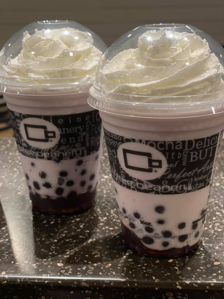 Purple colored bubble tea with whipped cream. The two drinks are in plastic to-go cups, with a large brown band with various coffee descriptors written on it. There circular logo is featured in the middle.