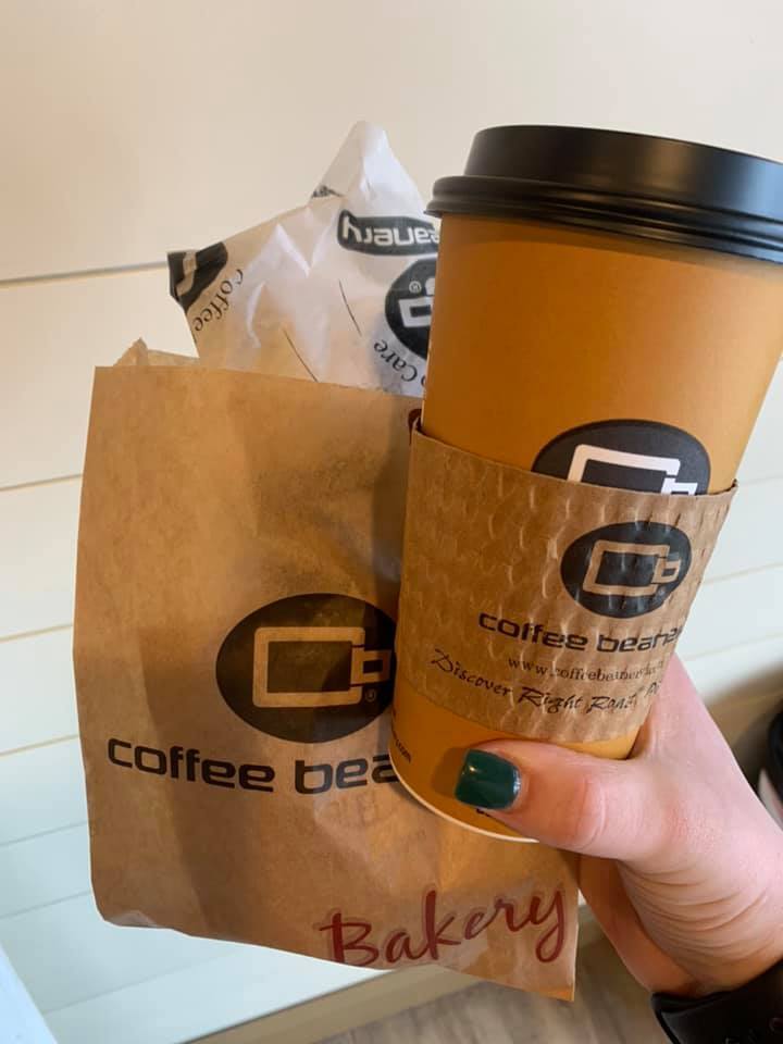 Hand holding a brown pastry bag, a white pastry bag, and a hot to-go coffee cup. All items feature a circular logo that contains a coffee cup.