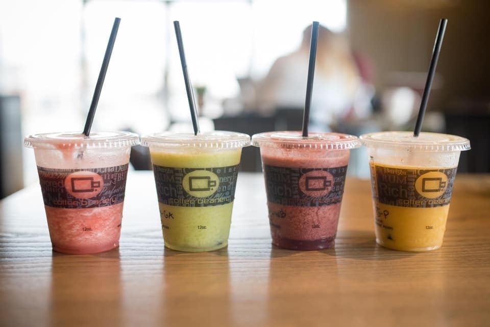 Four small smoothie drinks with black tall straws lined up in a row. From left to right the colors are light pink, green, a pink-purple, and orange.