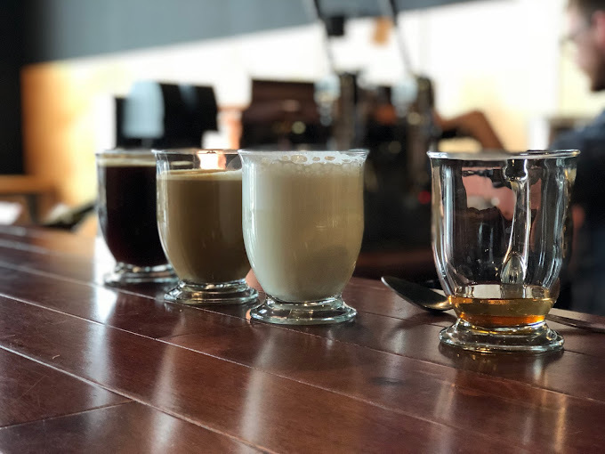 Various coffee stages ranging from light to dark, as well as a syrup, all in different glass cups. These cups are set on a dark wooden counter.