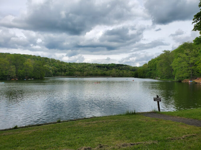 Lake view with surrounding trees in the background and grass in the foreground. There is a dirt path that goes close to the water, with a small unreadable sign beside it.