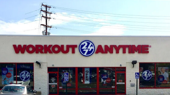 Front view of a flat beige building. The entrance and windows have red accenting. There is a big sign across the front that says "workout anytime" in red. A blue "24/7" circular logo sits between the two previous words.