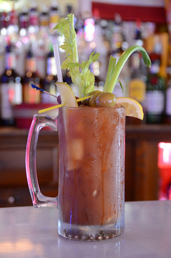 A view of a bloody Mary setting on a bar, loaded with celery, cheese, olives and pickles.