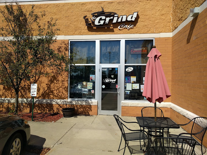 An outdoor view of an entrance to a coffee shop, the building is brick and has windows on either side of the glass door. A set of table and chairs with an umbrella sets out front of the doorway.