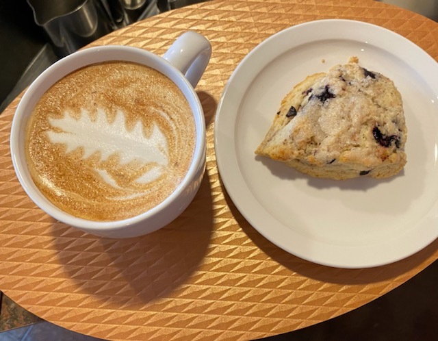A white plate consisting of a berry scone, paired with a latte with a flower latte art in it.