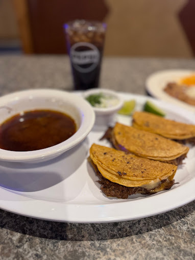 A white plate consisting of tacos and sauce to dip them in on the side in a white bowl.