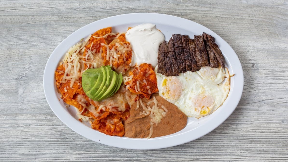 A white plate with a entre topped with sliced guac, a side of crema, beans, eggs, and a side of chorizo.