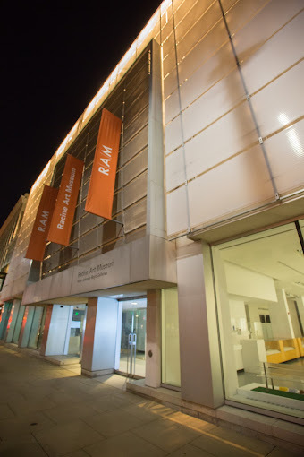 An outdoor view of a modern looking building, with plenty of large windows and red flags decorating the entrance to the building.