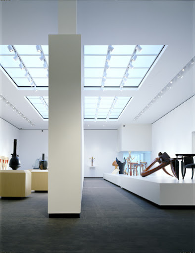An indoor view of a very large room with large sky lights illuminating the art pieces on large white pedestals below.