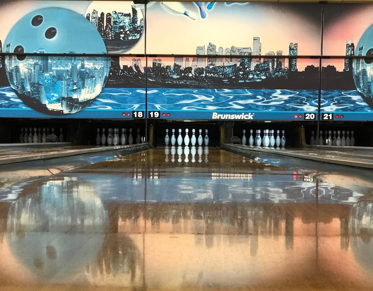A view of a bowling lane, a sunset with a city skyline is depicted on the top of the wall above the pins.