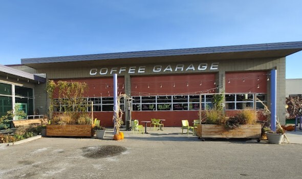 Grey and red garage type building. In front are plants in wooden crates and the parking lot.