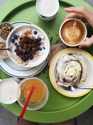 Overhead view of an orange iced drink, four hot coffees, a bowl, and a roll, all on a green dish.