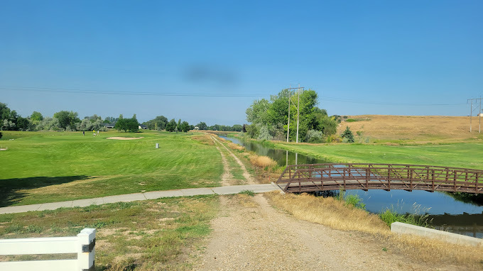 Green landscape. Dirt golf path lines the edge of the river that runs along the right side of the image. A concrete path leads to a bridge that crosses over the small river.