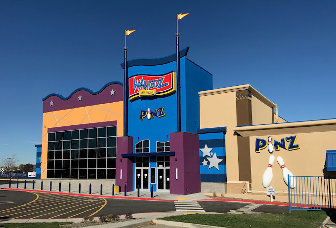 Exterior view of a blue, purple, orange, and beige whimsical building, Lining the entrance are two columns topped with yellow flags.
