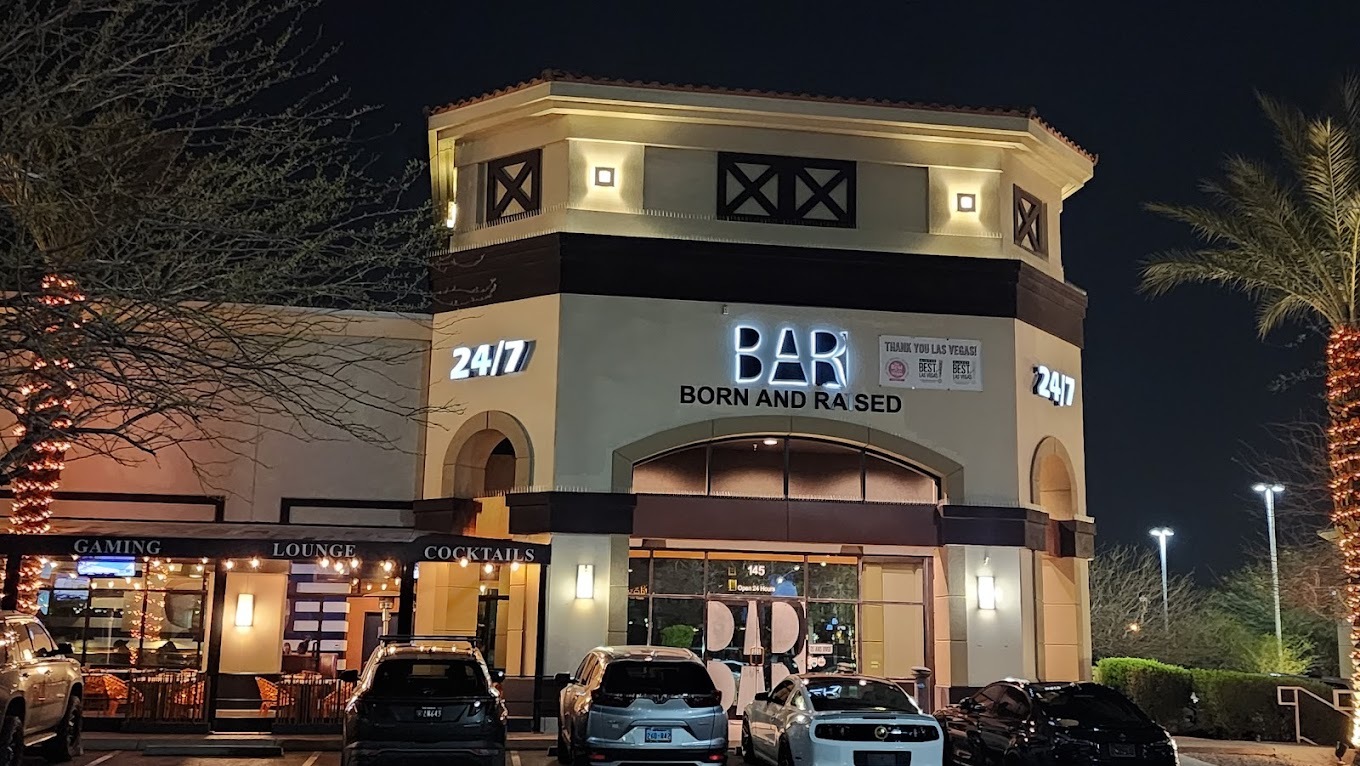 Outside view of a social bar that is beige, has rounded doorways, and a large white lighting sign that reads "24/7 Bar"