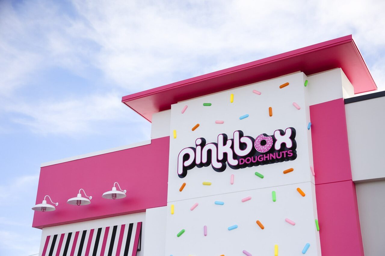 An outside view of a donut shop, a bright pink and white detailed with colorful sprinkle shaped lights, with a sign reading "Pinkbox Doughnuts"