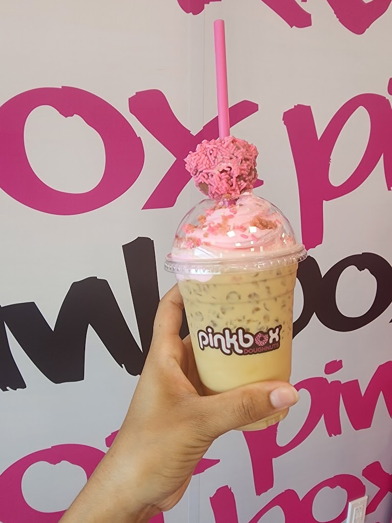 An iced coffee drink held up behind a background with pink and black lettering.