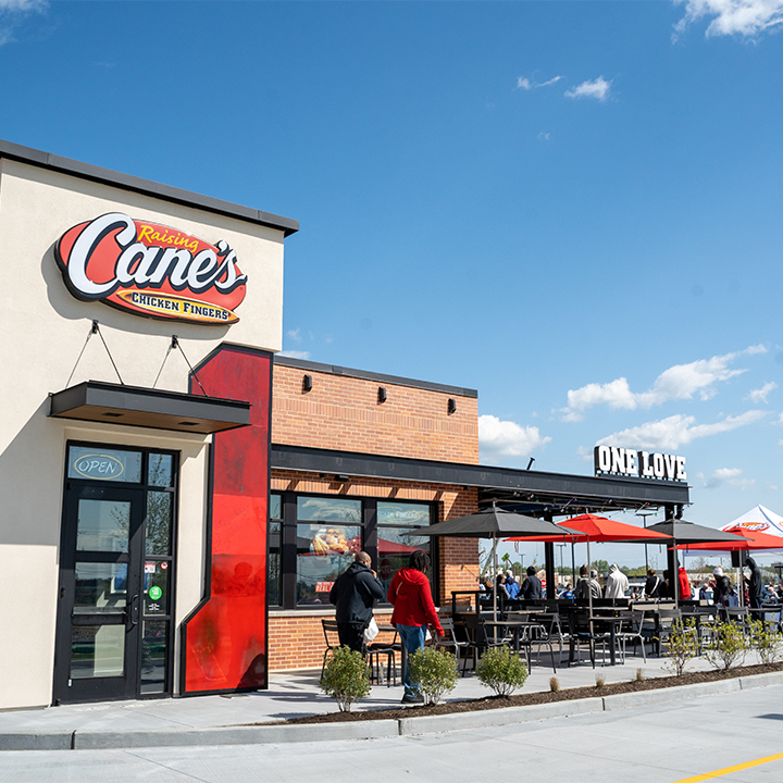 An outside view of a Raising Canes, white and brick with a small sliver of red accenting. A large red sign that reads "Raising Canes" is above the door way, outdoor seating is featured outside the front of the venue.