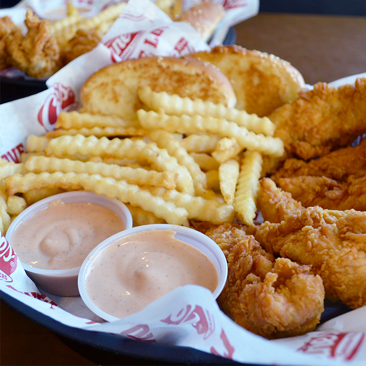 A close up view of a chicken tender basket meal, with a side of fries and Texas toast, as well as two sides of Cane's Sauce.