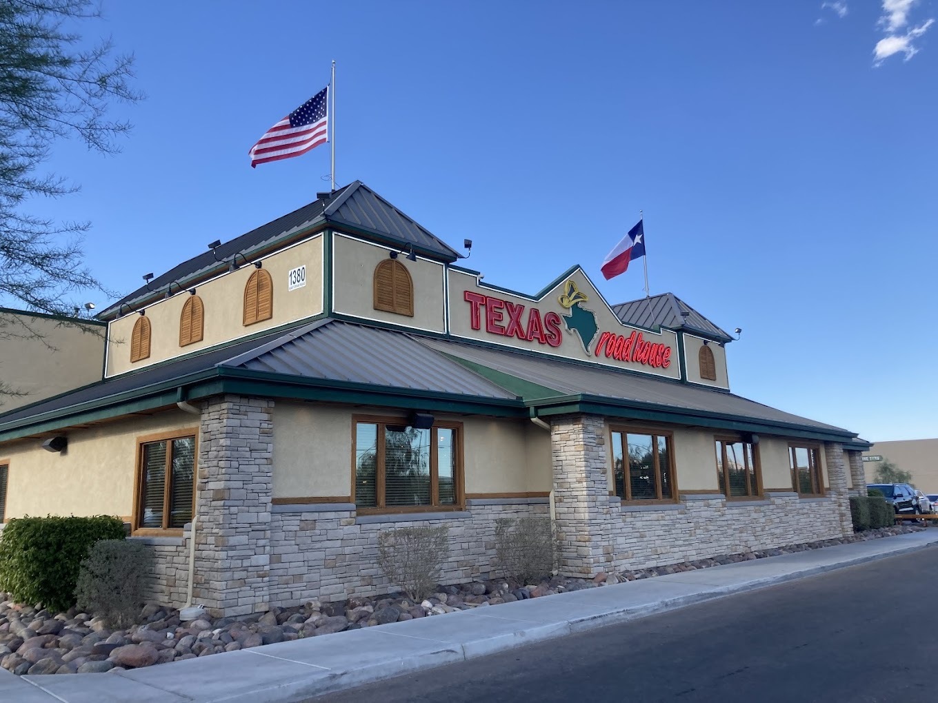An outside view of a Texas Roadhouse, a light beige colored building with red and green accenting, along with flags on the top of the building.