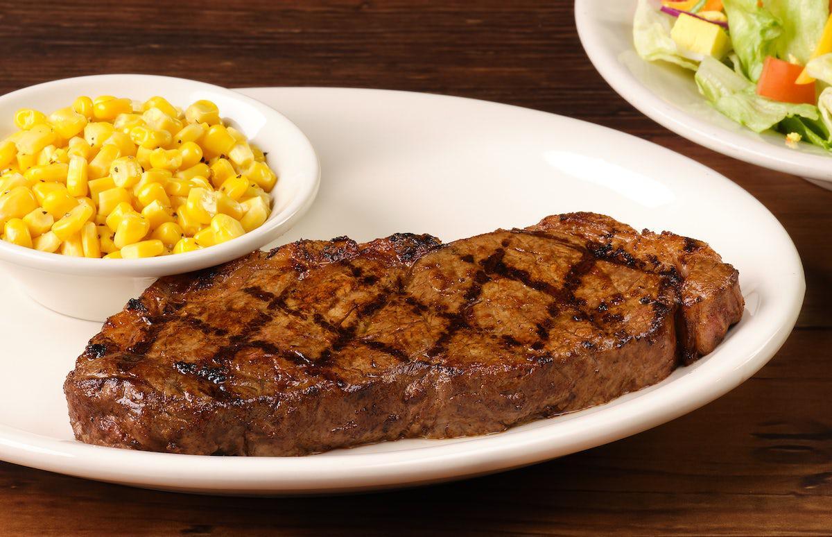 A white plate with a Texas Roadhouse steak, along with a side of corn in a white bowl.