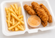 Crispy fried chicken tenders served with crinkle-cut fries and a small cup of dipping sauce in a white takeout container.