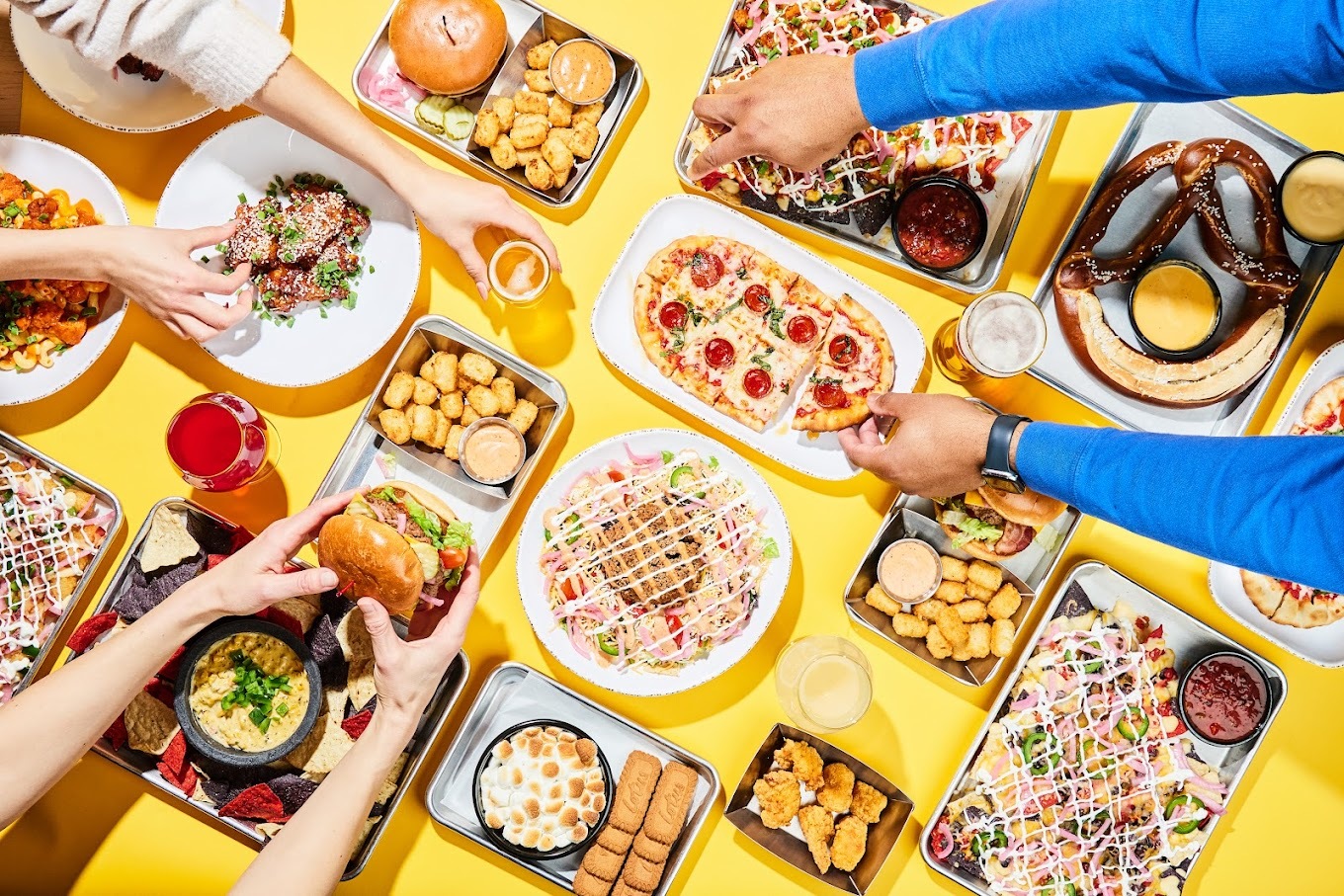 A white background with many plates of food like pizza, burgers, fries, with three sets of hands digging into the food.
