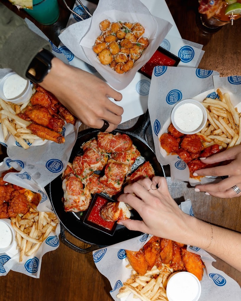 A table full of food trays with wings, French fries, and more. Two hands are picking around the food.