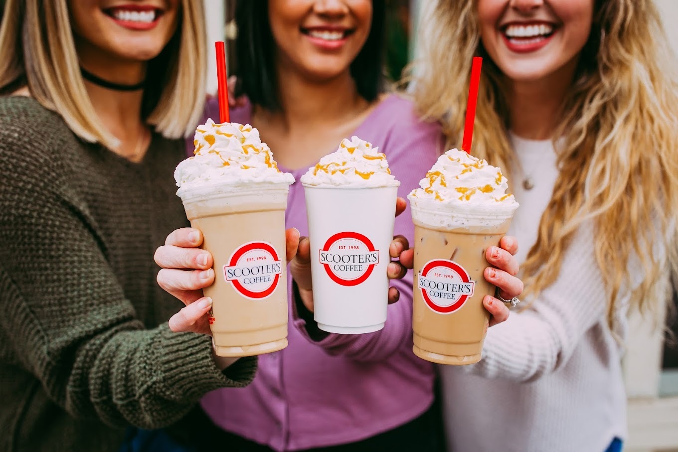 Three women are holding large frozen coffee drinks and holding them together as a group.