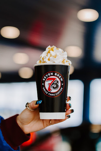 Hand with blue nails and long sleeve shirt holding a hot drink with whipped cream and caramel in a black cup.