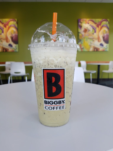Close view of a light iced coffee with whipped cream with an orange straw in a plastic cup. The logo on the cup is a red and black letter "B."