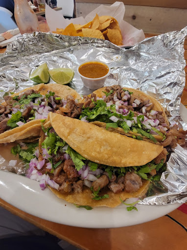 Three tacos spread on foil in a white plate. There are chips seen in the background.