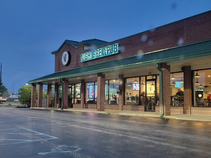 Exterior night view of a red brick building with columns and a green sunshade. There are two signs above the entrance, to the far left is a circular logo, and beside that is a green light up sign that states "Nigh Brewpub."