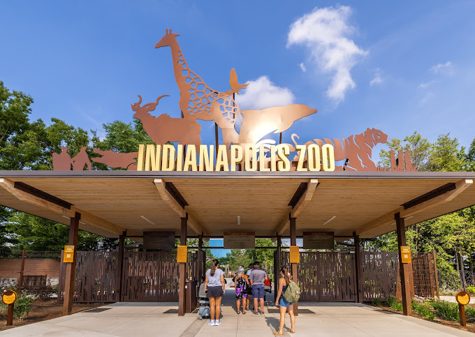 Entrance to Indianapolis Zoo with a large sunshade. Above the lettering are silhouette animals. The entrance has a fence and shows people walking through it.