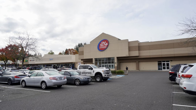 Exterior view of a flat brown building and a large parking lot with numerous cars. The section above the door entrance leads up to a triangular shape, with the circular red logo in the center.