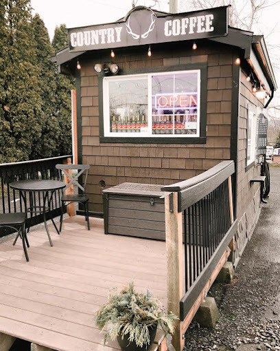 Small, quaint coffee stand with a small outdoor patio and fence. There are string lights hung around the roof of the building. There is a window by the back patio, and a sign above it that says "country coffee," with antlers in the middle.