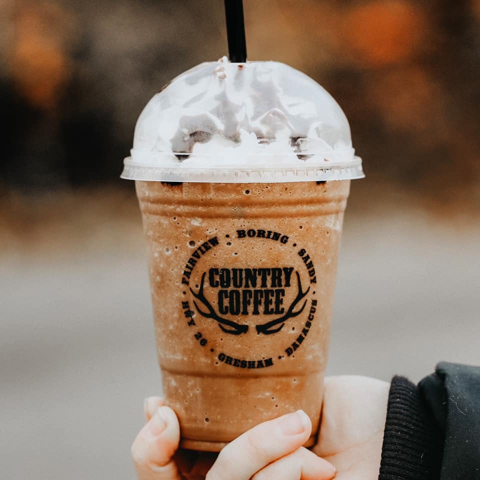 Blended iced coffee with whipped cream. The logo on the cup says "Country Coffee," with an image of antlers in the middle. It has a black straw and a person wearing long sleeves holds the cup from the bottom.