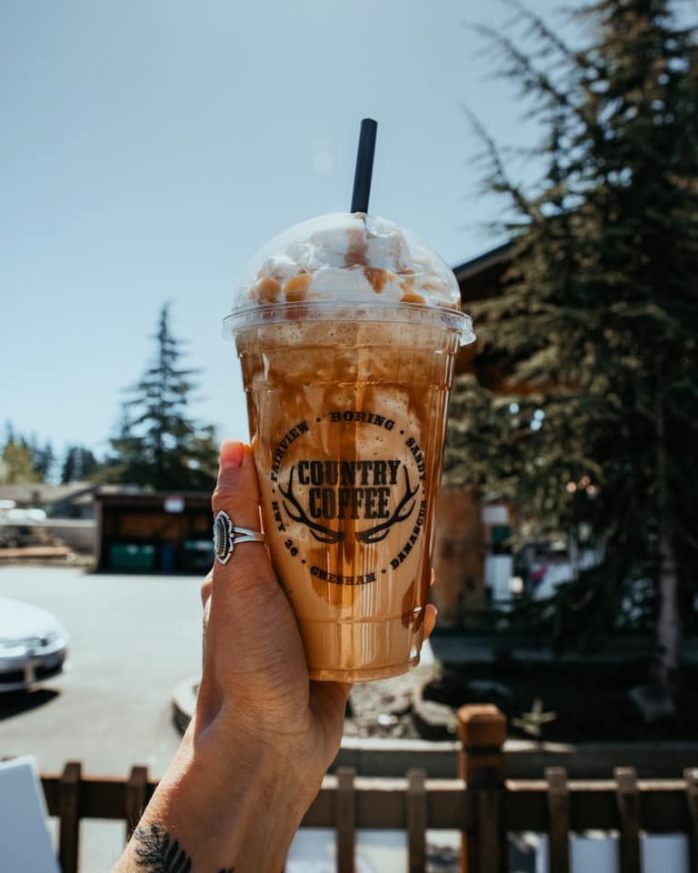 Hand holding a blended ice coffee with whipped cream and caramel. The background contains pine trees.