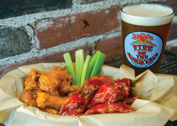 Wings with two different sauces, one orange and one red, in a yellow parchment paper basket. Off to the side is a glass cup filled with alcohol. The cup logo is blue and red.
