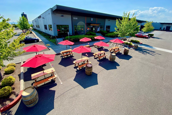 Exterior wide shot of a simplistic white and gray building. There are two rows of picnic seating with red umbrellas.