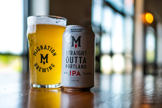 Glass filled with an alcoholic beverage next to a can of alcohol. They are set on a wooden table and have large windows in the background. Both beverages say "Migration Brewing," with the can additionally saying "Straight outta Portland."