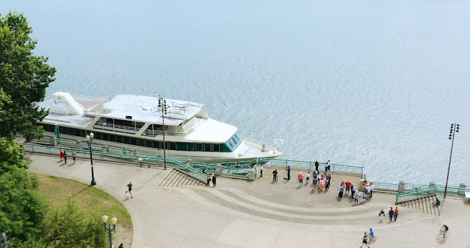 Aerial view of a docked cruise boat with people waiting to board. There is the water, sidewalk, and green fencing.