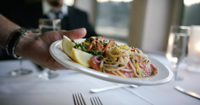 Hand holding a white plate containing spaghetti and sliced lemons. The background is a dining table with a white table cloth, silverware, and glasses.