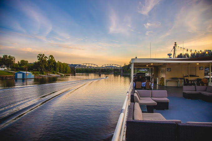 Sunset view of a river and the interior of a cruise boat. Along the river, there is a bridge going over it, as well as lots of trees near the edges.