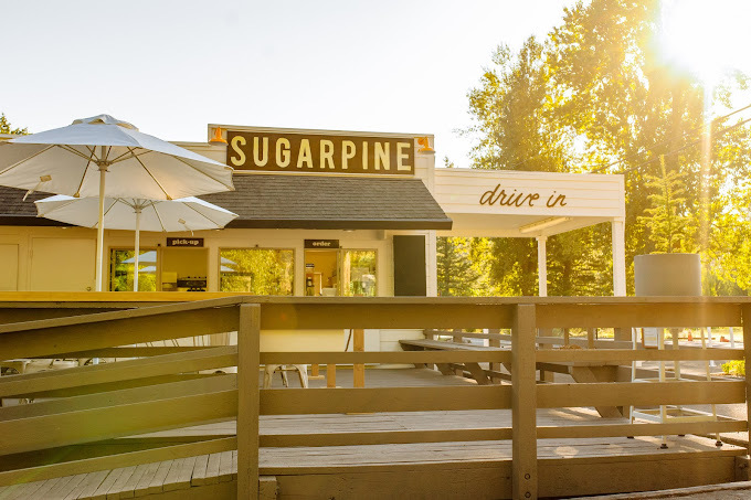 Sunny view of the front of the Sugarpine entrance. There is a walk up entrance with patio seating and two umbrellas, as well as a drive-in entrance on the side.